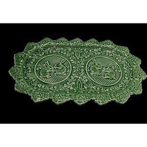 May include: An oval-shaped green ceramic tray featuring a scalloped edge with a raised floral pattern. The center displays two circular medallions, each containing a relief illustration of a rabbit surrounded by intricate vine and leaf details.