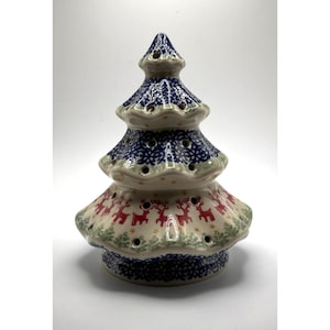 Puede incluir: Una figura de árbol de Navidad de cerámica con una base de color crema y patrones pintados a mano. Los niveles superiores muestran motivos florales azules, mientras que el nivel inferior presenta siluetas de renos rojos y pinos verdes con aberturas.