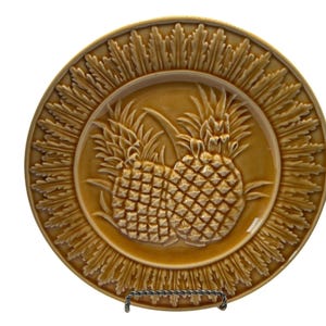 May include: A round ceramic plate in a warm golden-brown glaze featuring a central embossed design of two pineapples. The rim of the plate is decorated with a detailed, radiating leaf pattern that encircles the entire edge of the piece.