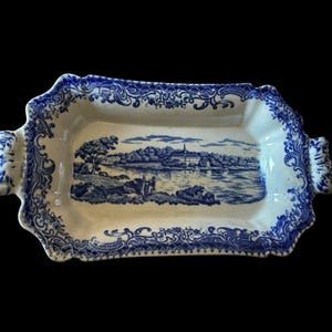 May include: A rectangular ceramic serving tray with handles, featuring a cream-colored base and a blue transferware pattern. The border displays intricate floral and scrollwork designs, while the center depicts a landscape with a building.