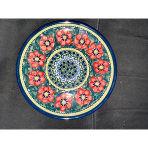 May include: A round ceramic plate featuring a central circular pattern of small blue squares surrounded by a ring of vibrant red flowers with yellow centers, set against a dark green leafy background with a yellow and blue border rim.