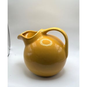 Może przedstawiać: Okrągły dzbanek ceramiczny w kolorze musztardowym o gładkim, błyszczącym wykończeniu. Naczynie posiada szeroki, pękaty korpus, zakrzywiony uchwyt przymocowany z boku oraz wyraźny, szeroki dzióbek na górnej krawędzi do nalewania płynów.