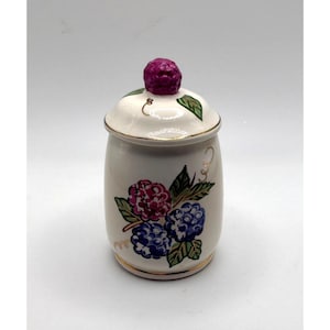May include: A cream-colored ceramic canister with a matching lid featuring a sculpted purple berry finial. The body and lid are decorated with hand-painted clusters of purple and blue berries, green leaves, and gold trim along the edges.