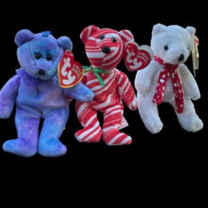 Può includere: Tre orsetti Ty Beanie Baby. Uno è blu con un fiocco blu, uno è a righe rosse e bianche e l'altro è bianco con una sciarpa rossa. Ogni orsetto ha un'etichetta rossa a forma di cuore.