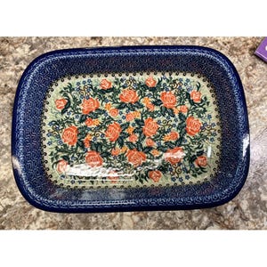 Könnte beinhalten: Eine rechteckige Auflaufform aus Keramik mit abgerundeten Ecken und einem dunkelblauen Rand mit geometrischem Muster. Das Innere zeigt ein florales Design mit orangefarbenen Rosen, grünen Blättern und kleinen blauen Blüten auf cremefarbenem Grund.