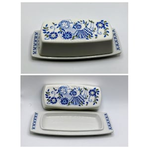 May include: A rectangular white ceramic butter dish featuring a blue and green floral pattern with a stylized figure on the lid. The base has a matching blue patterned border. The set includes a lid and a tray, shown both assembled and separated.