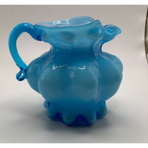 May include: A bright blue glass pitcher featuring a bulbous, rounded body with a ruffled, scalloped rim and a curved handle. The glass has a translucent quality with varying shades of blue, showcasing a smooth, polished surface and a flared base.