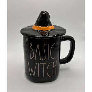 May include: A black ceramic mug with a matching lid shaped like a witch hat featuring an orange band. The mug has a handle on the right side and the text BASIC WITCH is written in a thin, light-colored, hand-lettered font on the front surface.