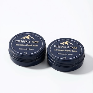 Puede incluir: Dos latas redondas de metal azul oscuro de Tussock & Tarn Kawakawa Repair Balm. Cada tapa presenta un gráfico de montaña dorado, el nombre de la marca, el nombre del producto, el texto Backcountry Ready y el peso de 30g, dentro de un círculo.