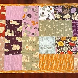 May include: A rectangular patchwork table runner featuring a variety of fabric squares with diverse patterns including floral, leaf, and geometric prints in shades of orange, brown, purple, pink, green, and blue, finished with a mustard yellow border.