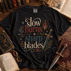 Può includere: T-shirt nera con la scritta "slow burns + sharp blades" in un carattere decorativo. Il design include una fiamma, una spada ed elementi floreali. Sono visibili libri e una collana.