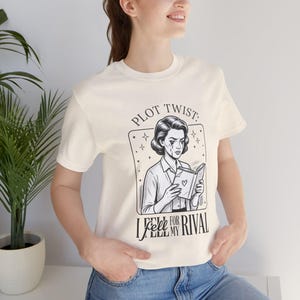 以下が含まれることがあります： クリーム色のTシャツ。白黒のイラストで、本を読む女性が描かれ、「PLOT TWIST: I FELL FOR MY RIVAL」の文字が入っています。デザインには星とハートが含まれています。Tシャツは半袖です。