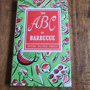 May include: A hardcover book titled "The ABC of Barbecue" by Peter Pauper Press. The cover features a red rectangle with white text, set against a green background with barbecue-themed illustrations in red and white. The book is on a wooden surface.