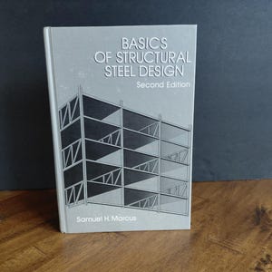 May include: A book titled "BASICS OF STRUCTURAL STEEL DESIGN, Second Edition" by Samuel H. Marcus. The cover is light gray with a black and white architectural illustration. The book is standing on a wooden surface.