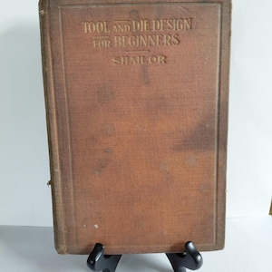 May include: A vintage hardcover book titled Tool and Die Design for Beginners by Shailor, featuring a weathered, textured brown fabric cover with embossed gold lettering. The book is displayed upright on a small, black plastic stand.