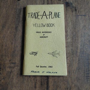 May include: A vintage "Trade-A-Plane Yellow Book" with the text "Price Reference of Aircraft." The cover is a light yellow color with black text and illustrations of airplanes. The book is dated Fall Quarter, 1964, and is signed by Frank J. Walker.