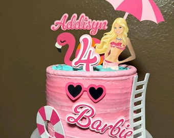 May include: A pink frosted cake decorated with a pool-themed topper set featuring a blonde doll, a pink flamingo, a pink and white umbrella, a white ladder, a pink and white life preserver, heart-shaped sunglasses, and text reading Addisyn 4 Barbie.