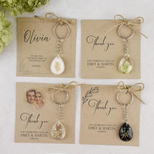 May include: Four keychains with teardrop-shaped pendants containing dried flowers or seeds. Each keychain is attached to a kraft paper card with a thank you message and names. The cards are tied with twine.
