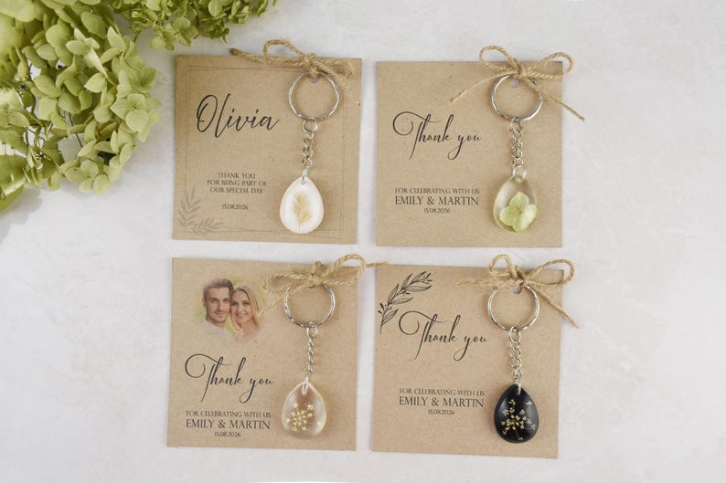 May include: Four keychains with teardrop-shaped pendants containing dried flowers or seeds. Each keychain is attached to a kraft paper card with a thank you message and names. The cards are tied with twine.
