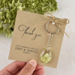 May include: A kraft paper card with the words "Thank you" in elegant script, and the names Emily & Martin with a date. A clear resin teardrop keychain with a dried flower is attached with a silver chain and ring.