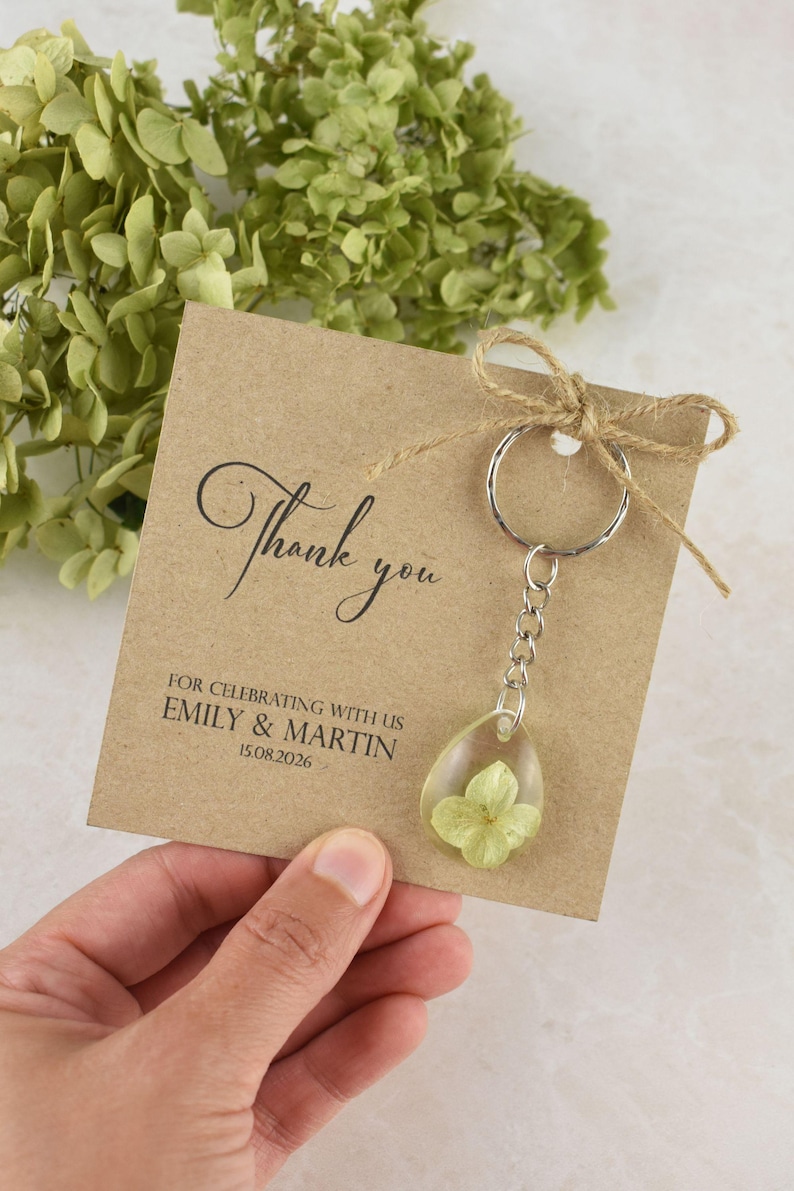 May include: A kraft paper card with the words "Thank you" in elegant script, and the names Emily & Martin with a date. A clear resin teardrop keychain with a dried flower is attached with a silver chain and ring.