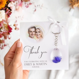 May include: A wedding favor featuring a clear resin teardrop-shaped keychain with a blue flower. The keychain hangs from a card with a photo of a couple and the words "Thank you" and wedding details.