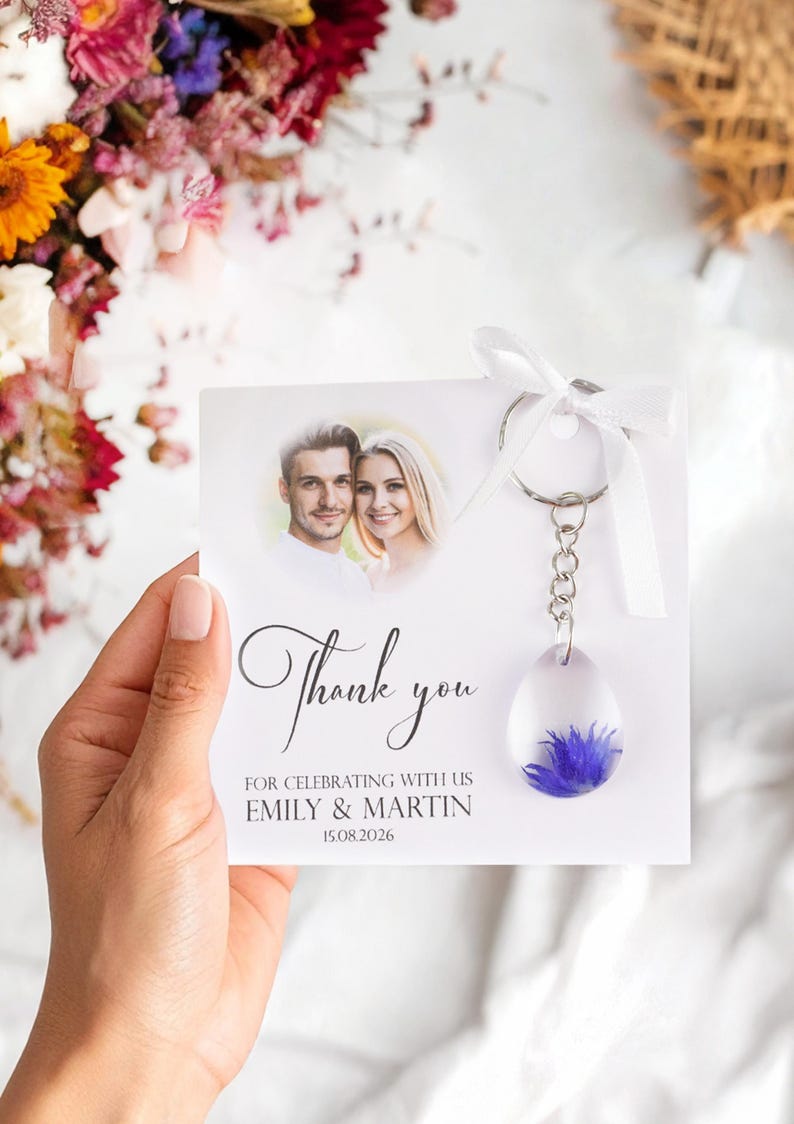 May include: A wedding favor featuring a clear resin teardrop-shaped keychain with a blue flower. The keychain hangs from a card with a photo of a couple and the words "Thank you" and wedding details.
