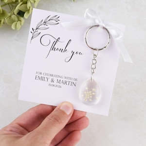 May include: A white card with the words "Thank you" in elegant script, accompanied by a keychain with a clear, teardrop-shaped charm filled with small, yellow flowers. The card also includes the names Emily & Martin and the date 15.08.2026.