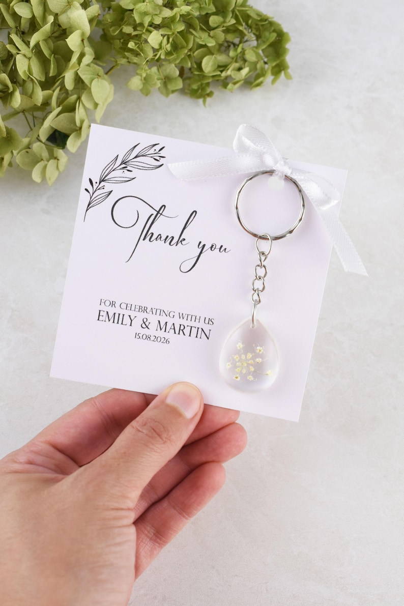 May include: A white card with the words "Thank you" in elegant script, accompanied by a keychain with a clear, teardrop-shaped charm filled with small, yellow flowers. The card also includes the names Emily & Martin and the date 15.08.2026.