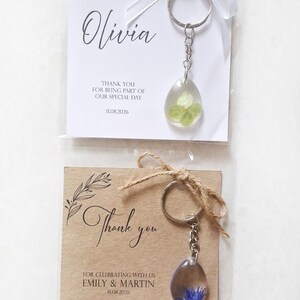 May include: Two keychains with resin pendants containing dried flowers. One keychain is paired with a white card that says "Olivia" and "Thank you for being part of our special day 15.08.2026". The other keychain is paired with a brown card that says "Thank you" and "For celebrating with us Emily & Martin 15.08.2026".