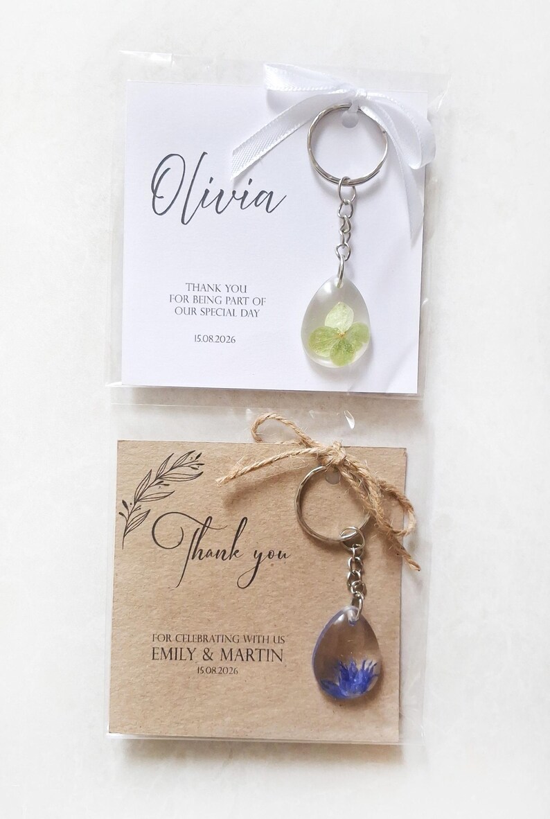 May include: Two keychains with resin pendants containing dried flowers. One keychain is paired with a white card that says "Olivia" and "Thank you for being part of our special day 15.08.2026". The other keychain is paired with a brown card that says "Thank you" and "For celebrating with us Emily & Martin 15.08.2026".