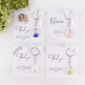 May include: Four keychains with cards, perfect for wedding favors. Each keychain has a clear, teardrop-shaped resin pendant with dried flowers. The cards say "Thank you" and include names and dates. Tied with white ribbons.