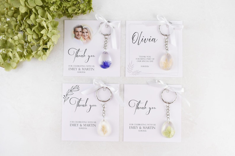 May include: Four keychains with cards, perfect for wedding favors. Each keychain has a clear, teardrop-shaped resin pendant with dried flowers. The cards say "Thank you" and include names and dates. Tied with white ribbons.