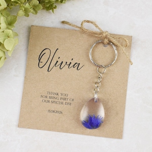 May include: A clear resin teardrop-shaped keychain with a blue flower embedded inside, attached to a silver keyring. The keychain is displayed with a brown card that reads "Olivia" and "Thank you for being part of our special day 15.08.2026".