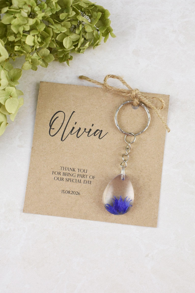 May include: A clear resin teardrop-shaped keychain with a blue flower embedded inside, attached to a silver keyring. The keychain is displayed with a brown card that reads "Olivia" and "Thank you for being part of our special day 15.08.2026".