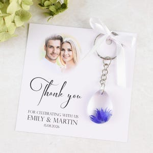 May include: A wedding favor featuring a clear, teardrop-shaped keychain with a blue flower encased inside. The keychain hangs from a card with a photo of a couple and the words "Thank you" and the names "Emily & Martin".