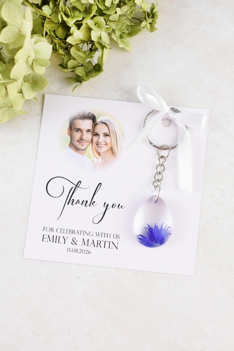 May include: A wedding favor featuring a clear, teardrop-shaped keychain with a blue flower encased inside. The keychain hangs from a card with a photo of a couple and the words "Thank you" and the names "Emily & Martin".
