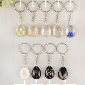 May include: A collection of nine keychains with teardrop-shaped resin pendants. Each keychain features a different dried flower or lace design, including hydrangea, bentgrass, and Queen Anne's Lace. The keychains have silver-colored metal rings and chains.