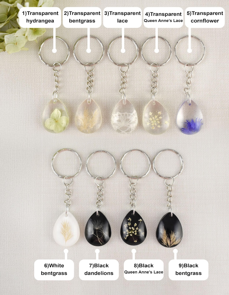 May include: A collection of nine keychains with teardrop-shaped resin pendants. Each keychain features a different dried flower or lace design, including hydrangea, bentgrass, and Queen Anne's Lace. The keychains have silver-colored metal rings and chains.