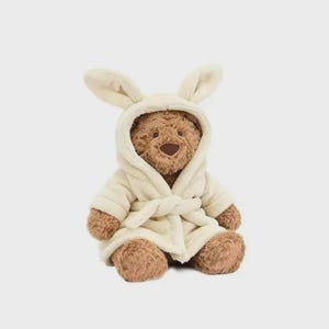Op de afbeelding: Een bruine teddybeer draagt een cr&egrave;mekleurige badjas met een capuchon en konijnenoren. De badjas is in de taille vastgebonden met een bijpassende riem. De beer heeft een bruine neus en poten.
