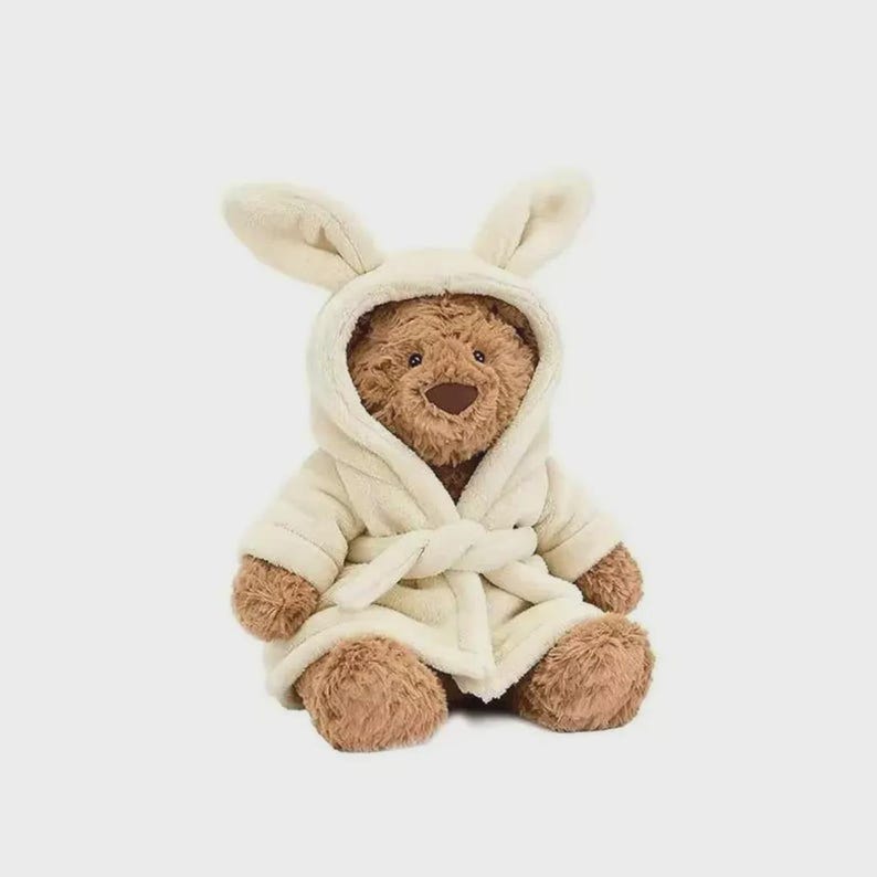 Op de afbeelding: Een bruine teddybeer draagt een cr&egrave;mekleurige badjas met een capuchon en konijnenoren. De badjas is in de taille vastgebonden met een bijpassende riem. De beer heeft een bruine neus en poten.