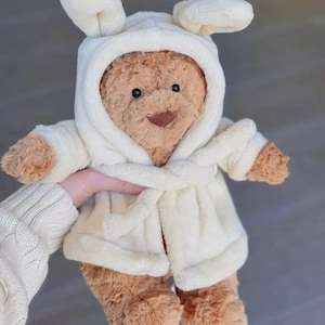 Op de afbeelding: Een bruine teddybeer met een witte badjas met capuchon en konijnenoren. De beer heeft een lachend gezicht en houdt zijn armen uit. De badjas is in de taille vastgebonden met een bijpassende riem.
