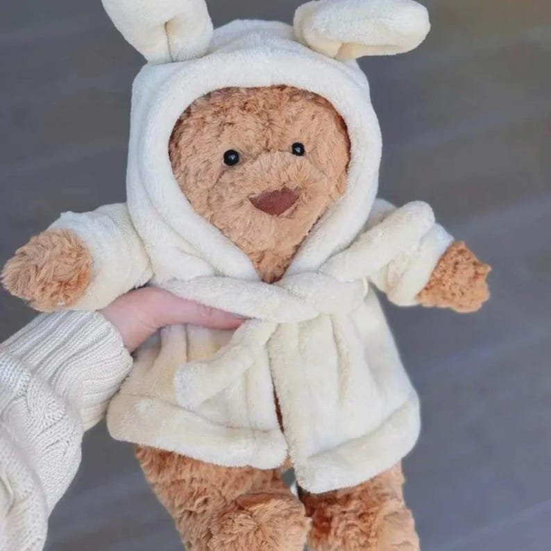 Op de afbeelding: Een bruine teddybeer met een witte badjas met capuchon en konijnenoren. De beer heeft een lachend gezicht en houdt zijn armen uit. De badjas is in de taille vastgebonden met een bijpassende riem.