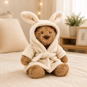 Op de afbeelding: Een bruine teddybeer met een cr&egrave;mekleurige badjas met capuchon en konijnenoren. De badjas is in de taille vastgebonden met een bijpassende riem. De beer zit op een cr&egrave;mekleurig sprei.