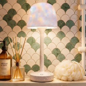 May include: A white mushroom-shaped table lamp with a soft, multi-colored glowing dome shade. The lamp features a slender stem and a circular base, both covered in small water droplets, standing next to a lotion bottle and a bath sponge.