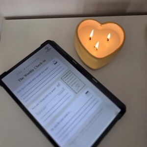 May include: A tablet displaying a digital document titled The Weekly Check-in with lines for writing, positioned next to a yellow heart-shaped candle with three lit wicks that emit a warm glow, creating a soft light on the surrounding surface.