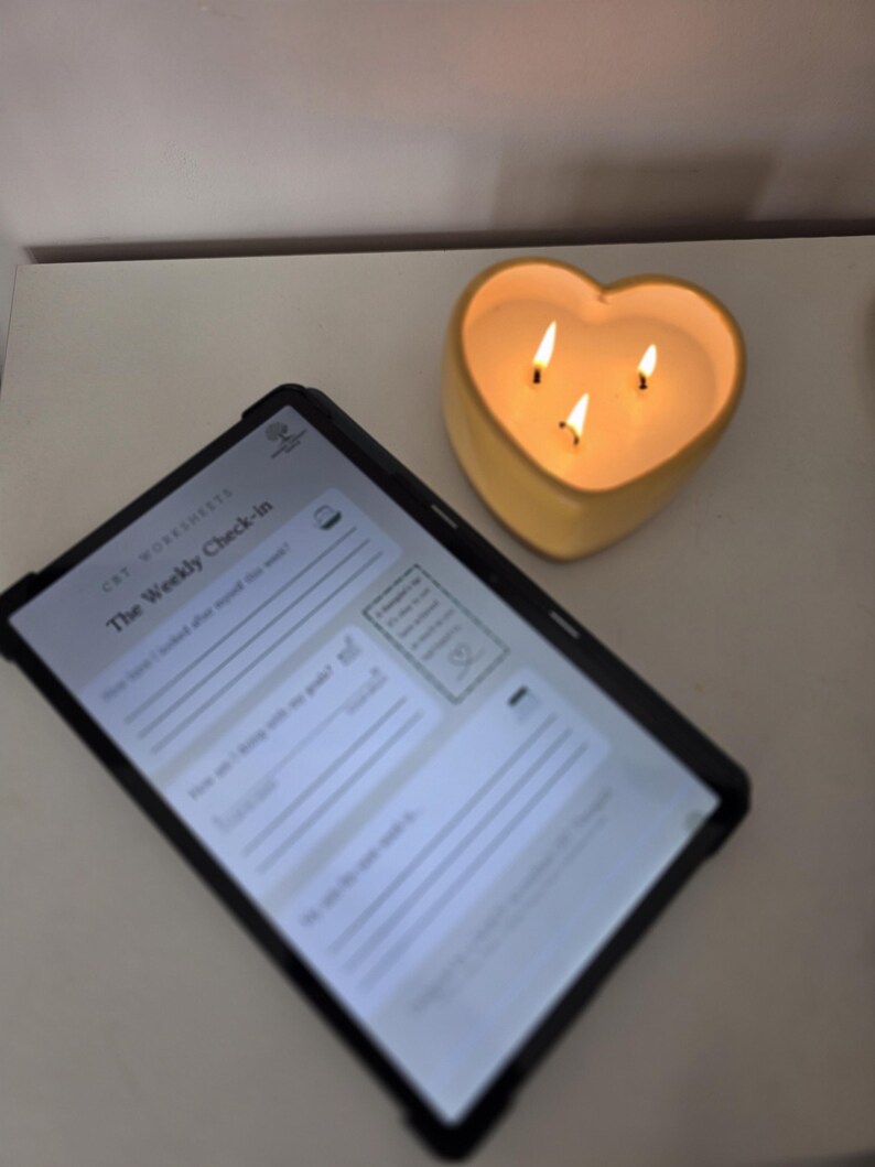 May include: A tablet displaying a digital document titled The Weekly Check-in with lines for writing, positioned next to a yellow heart-shaped candle with three lit wicks that emit a warm glow, creating a soft light on the surrounding surface.