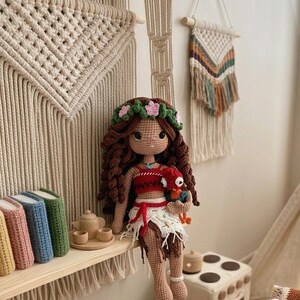 May include: A handmade crochet doll with long brown hair, a floral crown, and a red and white outfit. The doll is sitting on a shelf with books and a tea set. A basket filled with crochet toys and wooden blocks sits nearby.