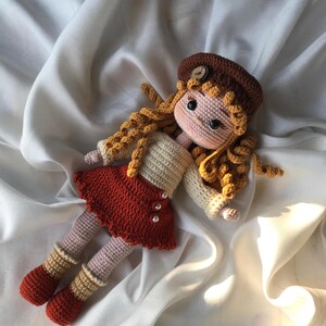 May include: A handmade crochet doll with curly blonde hair, wearing a brown beret, a cream-colored sweater, and a red skirt. The doll has pink legs and orange and cream striped socks. The doll is lying on a white satin surface.