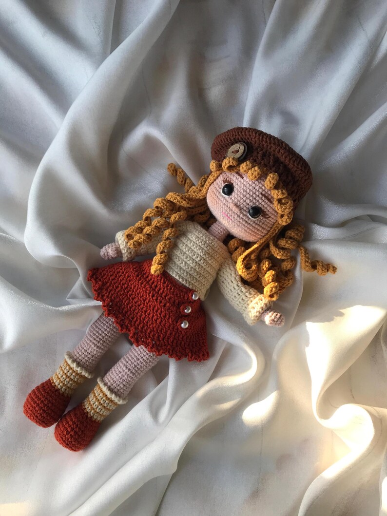 May include: A handmade crochet doll with curly blonde hair, wearing a brown beret, a cream-colored sweater, and a red skirt. The doll has pink legs and orange and cream striped socks. The doll is lying on a white satin surface.